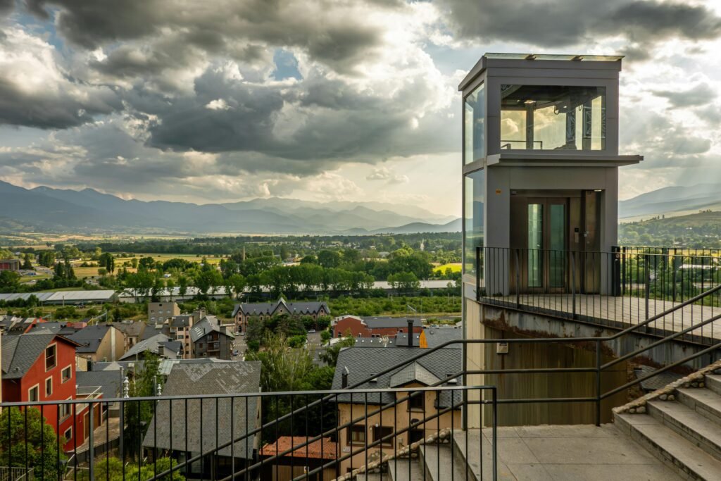 project-1 A modern elevator overlooks a picturesque town with mountains in the background under dramatic skies.